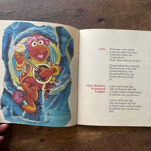 May include: An open book featuring a colorful illustration of a pink-furred Fraggle character playing a banjo on the left page. The right page contains printed lyrics titled Gobo and Gobo, Wembley & Gathered Fraggles with black text on cream paper.