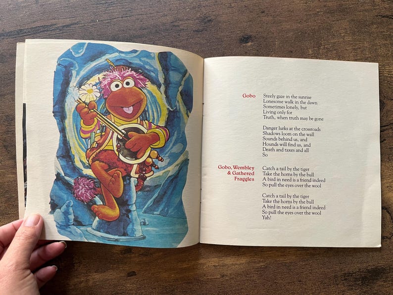 May include: An open book featuring a colorful illustration of a pink-furred Fraggle character playing a banjo on the left page. The right page contains printed lyrics titled Gobo and Gobo, Wembley & Gathered Fraggles with black text on cream paper.