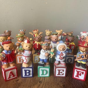 May include: A collection of twenty-six miniature bear figurines, each standing on a colorful alphabet block. The bears are dressed in various costumes, including a painter, a baseball player, a doctor, a winter explorer, and a firefighter.