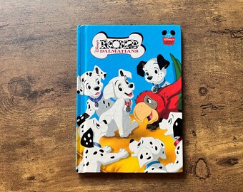 Pode incluir: Um livro infantil intitulado "102 Dálmatas" com uma capa azul. A capa apresenta ilustrações de vários filhotes dálmatas, um papagaio vermelho e o logotipo da Disney. O livro está sobre um fundo de madeira.
