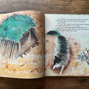 May include: An open children's storybook featuring a watercolor illustration of a wolf and a brush hut on the right page, with text that reads: Ma'ii rolled over and went back to sleep. But no sooner was he asleep again than the faraway voice returned.