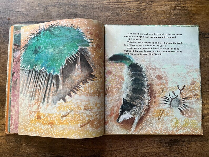 May include: An open children's storybook featuring a watercolor illustration of a wolf and a brush hut on the right page, with text that reads: Ma'ii rolled over and went back to sleep. But no sooner was he asleep again than the faraway voice returned.