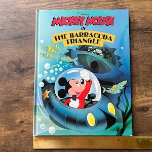 Op de afbeelding: Een hardcover boek getiteld Disney's Mickey Mouse in The Barracuda Triangle, met een illustratie van Mickey Mouse in een blauwe onderzee&euml;r. Een gele liniaal ligt onderaan en toont een breedte van ongeveer 21,5 cm.