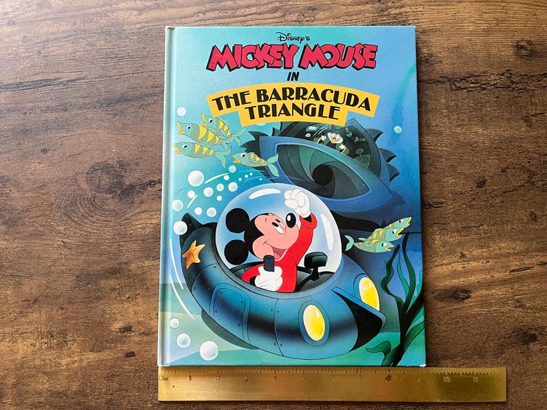 Op de afbeelding: Een hardcover boek getiteld Disney's Mickey Mouse in The Barracuda Triangle, met een illustratie van Mickey Mouse in een blauwe onderzee&euml;r. Een gele liniaal ligt onderaan en toont een breedte van ongeveer 21,5 cm.