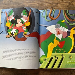 Op de afbeelding: Een open kinderboek met een kleurrijke illustratie van Mickey Mouse in een rood shirt en blauwe broek, bewaakt door twee in het groen geklede figuren. Op de rechterpagina bedient een groenhuidig personage met een cape een bedieningspaneel.
