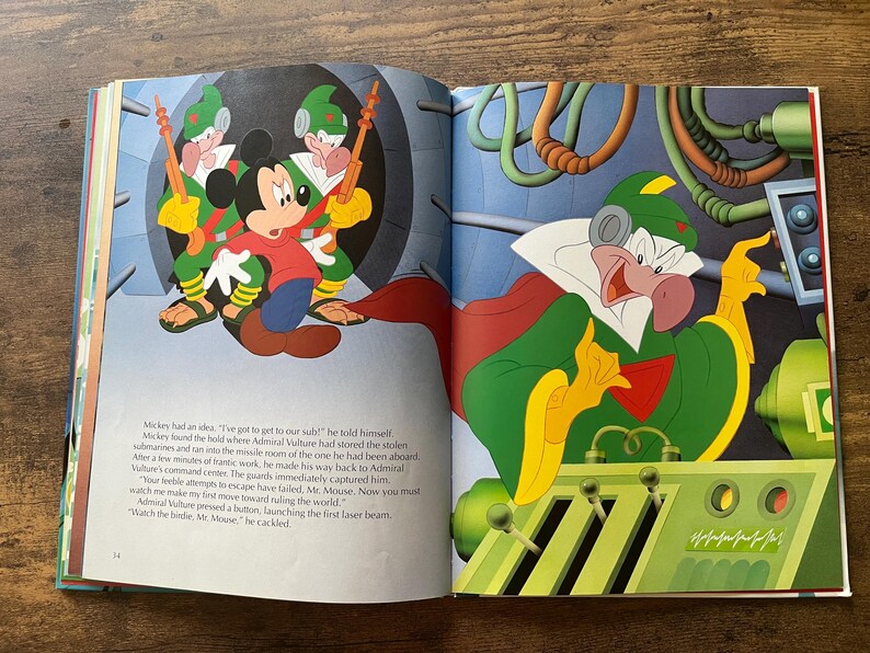 Op de afbeelding: Een open kinderboek met een kleurrijke illustratie van Mickey Mouse in een rood shirt en blauwe broek, bewaakt door twee in het groen geklede figuren. Op de rechterpagina bedient een groenhuidig personage met een cape een bedieningspaneel.