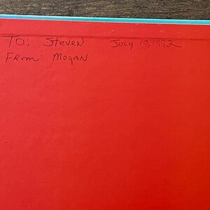 Op de afbeelding: Een felrode pagina in een boek met een lichtblauwe rand. Handgeschreven tekst in zwarte inkt luidt: To: Steven, From: Megan, July 10, 1992. Het oppervlak is glad en vlak en toont een duidelijke inscriptie op de levendige rode achtergrond.