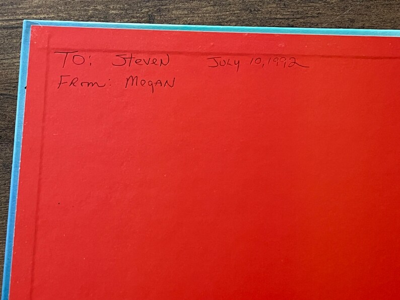 Op de afbeelding: Een felrode pagina in een boek met een lichtblauwe rand. Handgeschreven tekst in zwarte inkt luidt: To: Steven, From: Megan, July 10, 1992. Het oppervlak is glad en vlak en toont een duidelijke inscriptie op de levendige rode achtergrond.