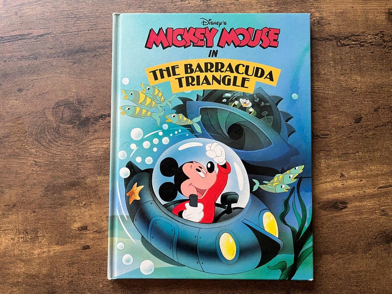 May include: A hardcover book titled Disney's Mickey Mouse in The Barracuda Triangle. The cover features a colorful illustration of Mickey Mouse inside a blue submarine, surrounded by green fish, bubbles, and a dark underwater structure.