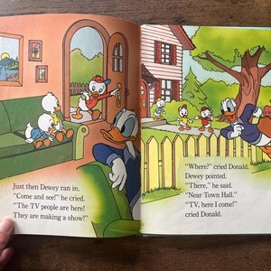 May include: An open children's storybook featuring colorful illustrations of Donald Duck and his nephews. The left page shows characters in a living room, while the right page depicts them outdoors near a house. Text accompanies the illustrations.