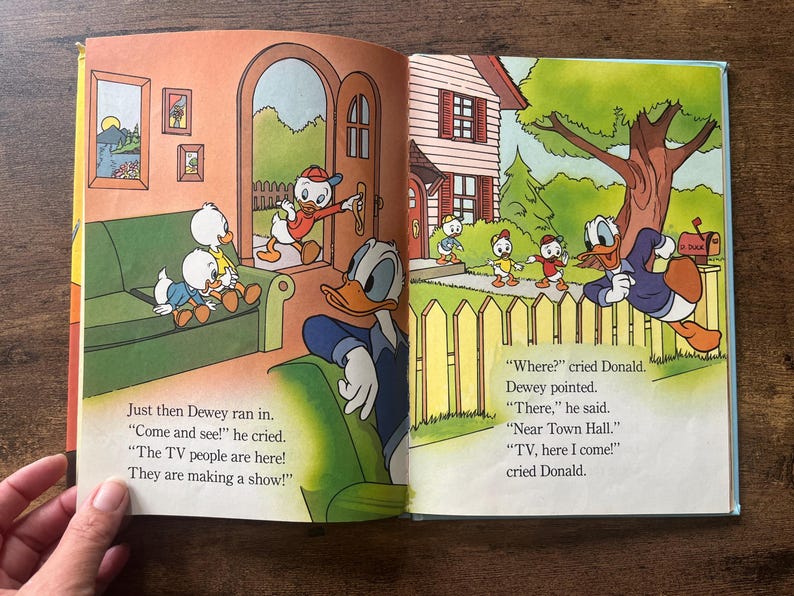 May include: An open children's storybook featuring colorful illustrations of Donald Duck and his nephews. The left page shows characters in a living room, while the right page depicts them outdoors near a house. Text accompanies the illustrations.