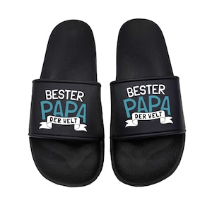 Könnte beinhalten: Ein Paar schwarze Schlappen mit dem Text "BESTER PAPA DER WELT" in weißer und blauer Schrift.