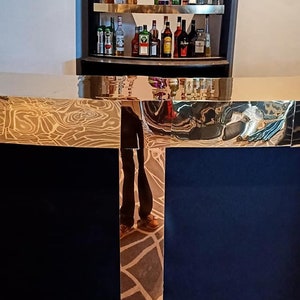 May include: A gold-trimmed, black bar counter with a mirrored surface reflecting a person and a shelf of liquor bottles. The shelf is made of wood and has a gold trim. The bottles are arranged in rows and are mostly clear glass with labels.