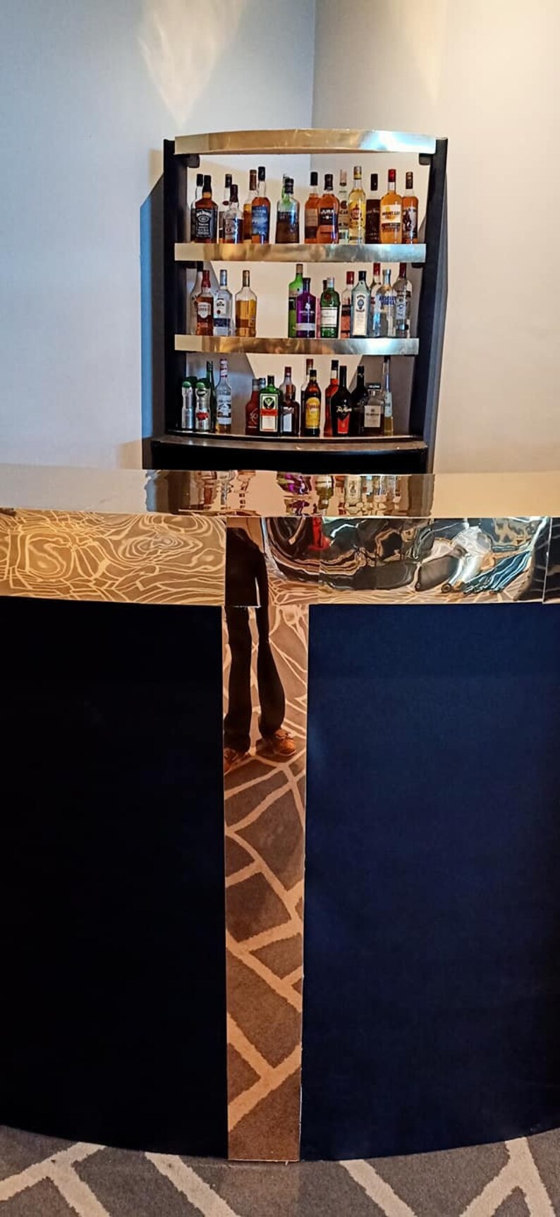May include: A gold-trimmed, black bar counter with a mirrored surface reflecting a person and a shelf of liquor bottles. The shelf is made of wood and has a gold trim. The bottles are arranged in rows and are mostly clear glass with labels.