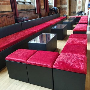 May include: A set of black velvet modular seating with red cushions. The seating is arranged in a long row with a few smaller square cushions in front. The seating is in a large room with wooden floors and red curtains.