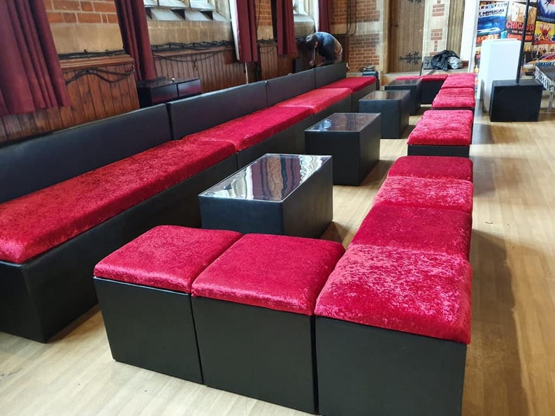 May include: A set of black velvet modular seating with red cushions. The seating is arranged in a long row with a few smaller square cushions in front. The seating is in a large room with wooden floors and red curtains.