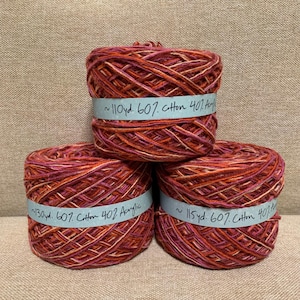 May include: Three skeins of yarn in shades of red, orange, and cream are stacked. Each skein has a light blue band with text indicating the yardage and fiber content: 60% cotton and 40% acrylic.