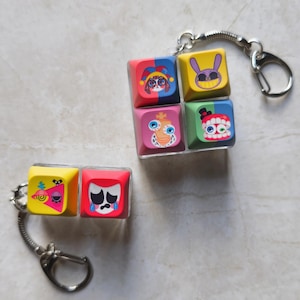 May include: Two keycap keychains featuring colourful graphic designs. One keychain has four square keycaps arranged in a grid, while the other has two side-by-side. The keycaps display various cartoon-style faces, including a jester, a rabbit, a crown-wearing figure, a toothy mouth, a pink triangle with an eye, and a sad face with blue tears.