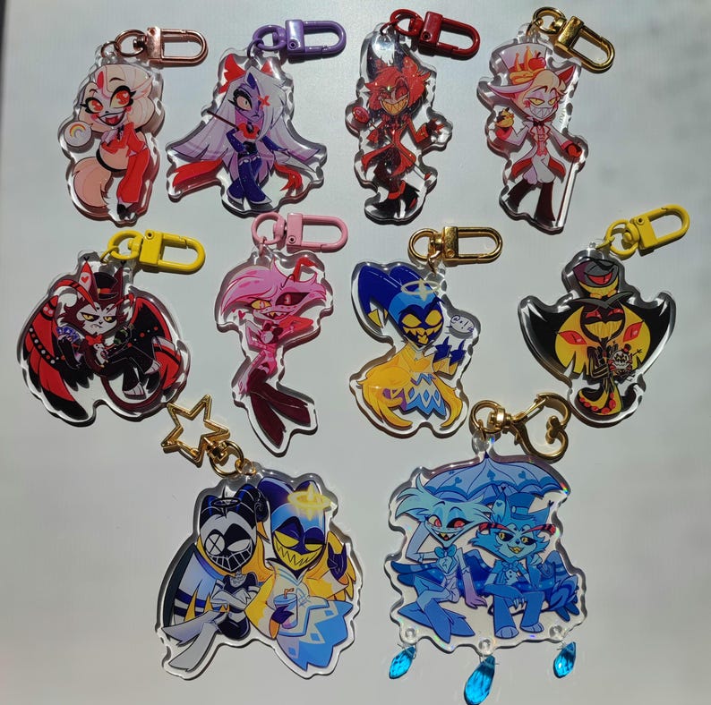 May include: A set of 10 colorful cartoon character keychains. The characters are from the animated series "Hazbin Hotel". The keychains are made of clear acrylic and feature different characters from the show, including Angel Dust, Alastor, and Charlie.