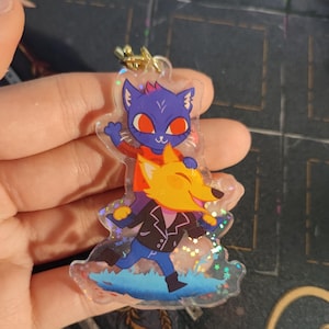 May include: A clear acrylic keychain featuring a blue cat character with red eyes and a yellow fox character with a white smile. The characters are standing on a blue and white background with glitter accents.
