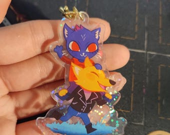 May include: A clear acrylic keychain featuring a blue cat character with red eyes and a yellow fox character with a white smile. The characters are standing on a blue and white background with glitter accents.