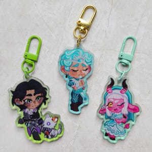 Può includere: Tre portachiavi in acrilico con personaggi illustrati. Uno mostra una figura dai capelli scuri con una piccola creatura bianca, uno una figura dai capelli blu con gli occhi chiusi e uno un personaggio dalla pelle rosa con corna e vestito.
