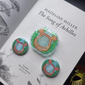 May include: Three decorative pins and a larger, clear, circular object with a lyre design and the text "He is half of my soul as the poets say." The objects are on a book page titled "The Song of Achilles."