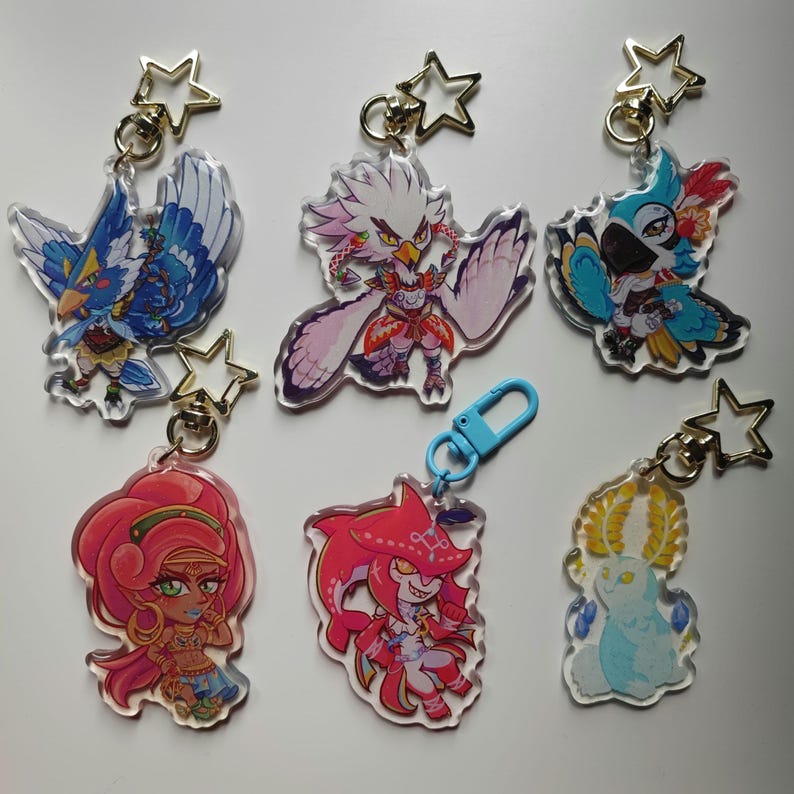 May include: Six colorful character keychains with gold star charms. The characters include a blue bird, a white bird, a blue parrot, a red-haired woman, a red fish, and a white creature with horns.