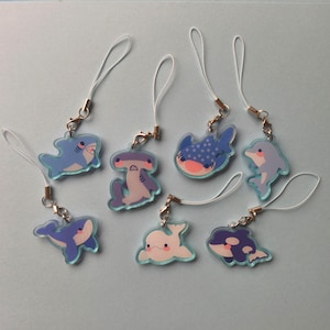 Peut inclure: Une collection de breloques d'animaux marins, dont un requin, un requin marteau, un requin-baleine, un dauphin, un béluga et une orque. Chaque breloque est d'une couleur différente et possède un cordon blanc attaché avec un fermoir doré.