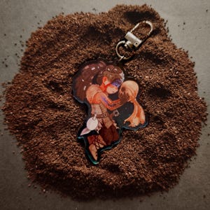 May include: A clear acrylic keychain featuring a stylized illustration of a person holding a skull. The keychain has a silver clasp and a small, decorative charm. The artwork is set against a dark background.