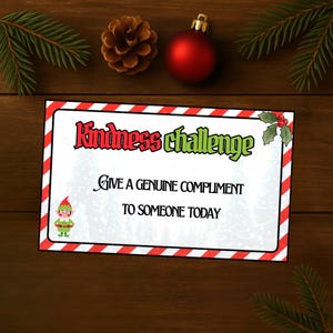 Op de afbeelding: Een kerstkaart met de tekst "Kindness Challenge" in rood en groen. De kaart moedigt aan om een oprecht compliment te geven. Een kleine elf-illustratie staat linksonder. De kaart ligt op een houten oppervlak met dennennaalden, een dennenappel en een rode kerstbal.
