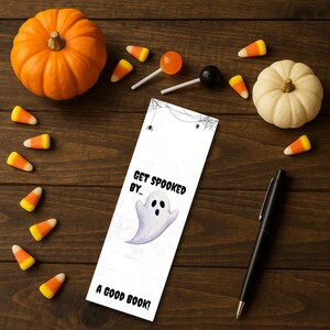 Op de afbeelding: Een Halloween-thema bladwijzer met een spook illustratie en de tekst "Get Spooked By... A Good Book!" is omringd door snoepmaïs, pompoenen en lolly's op een houten oppervlak. Er is ook een zwarte pen aanwezig.