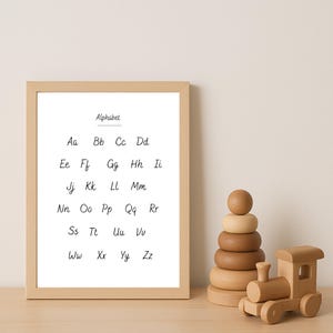 Op de afbeelding: Een ingelijste alfabetprint in een licht houten frame. De print toont de letters Aa tot en met Zz in een eenvoudig lettertype. Een houten stapelspeelgoed en een houten trein staan naast het frame.