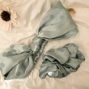 May include: Two sage green satin hair scrunchies with white polka dots. One scrunchie is tied in a bow shape, the other is a simple scrunchie.