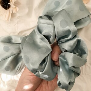 May include: A light blue satin scrunchie with white polka dots. The scrunchie is tied in a bow and is being held by a hand.