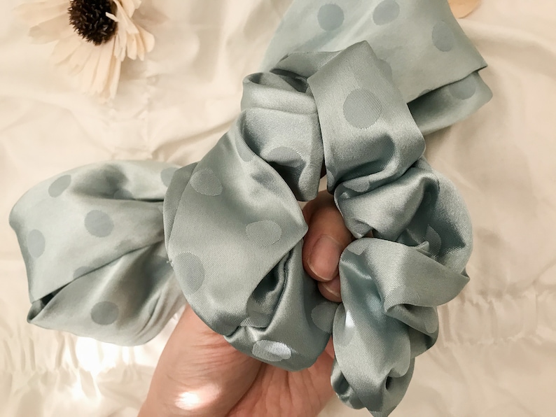May include: A light blue satin scrunchie with white polka dots. The scrunchie is tied in a bow and is being held by a hand.