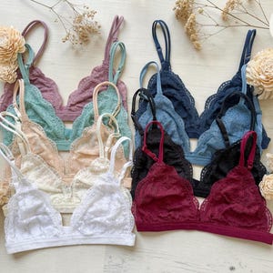 May include: A collection of lace bralettes in various colors, including white, cream, peach, teal, purple, navy, black, and burgundy. Each bralette features delicate lace detailing and adjustable straps. The bralettes are arranged on a light-colored surface, with dried flowers.