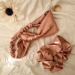 May include: A set of two satin hair accessories in a dusty rose color. The headband is tied in a knot and the scrunchie is gathered in a ball shape.