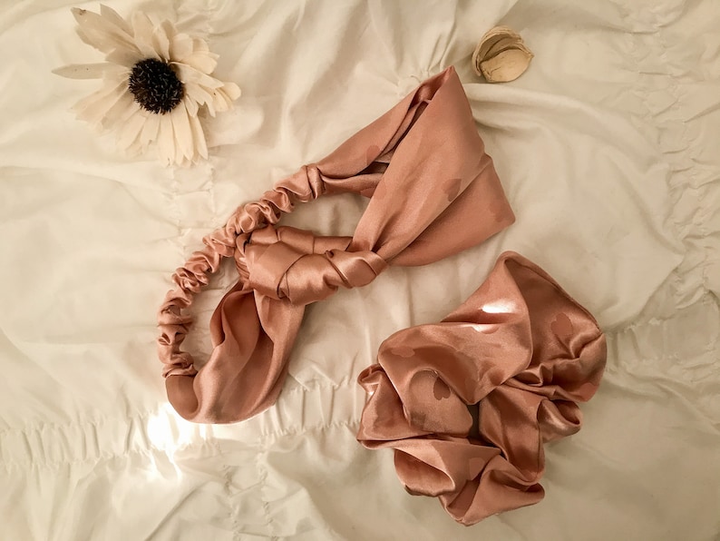 May include: A set of two satin hair accessories in a dusty rose color. The headband is tied in a knot and the scrunchie is gathered in a ball shape.