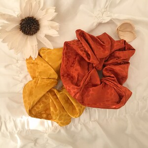 May include: Two fabric scrunchies, one orange and one yellow, with a white floral pattern. The scrunchies are on a white surface.