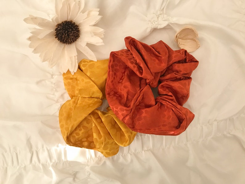 May include: Two fabric scrunchies, one orange and one yellow, with a white floral pattern. The scrunchies are on a white surface.