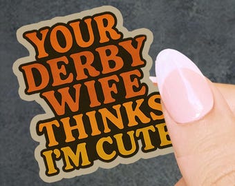 May include: A sticker with the text "YOUR DERBY WIFE THINKS I'M CUTE" in orange and yellow lettering. The sticker has a beige border and is held by a hand with a pink manicure.