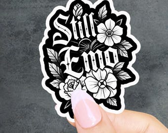 May include: A black and white sticker featuring the text Still Emo in a gothic-style font, surrounded by detailed illustrations of various flowers and leaves. The design is rendered in high-contrast black ink on a white background with a thick border.