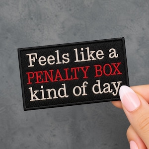 May include: Black rectangular patch with white and red embroidered text that reads "Feels like a PENALTY BOX kind of day". The patch is held by a person with a gray background.