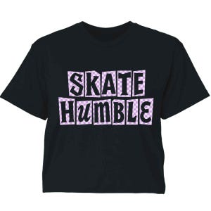 May include: A black short-sleeve t-shirt featuring the text "SKATE HUMBLE" in bold black letters. Each letter is set against an individual light purple and white checkered square background, creating a two-line graphic design on the chest.
