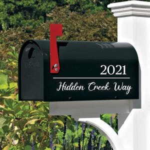 Puede incluir: Un buzón negro con una bandera roja, montado en un poste blanco. El texto "2021 Hidden Creek Way" se muestra en escritura blanca en el buzón. El fondo presenta follaje verde.