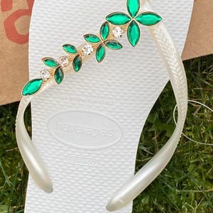 May include: White Havaianas flip-flop with a decorative strap featuring green and clear gemstones in a floral design. The flip-flop is on a grassy surface.