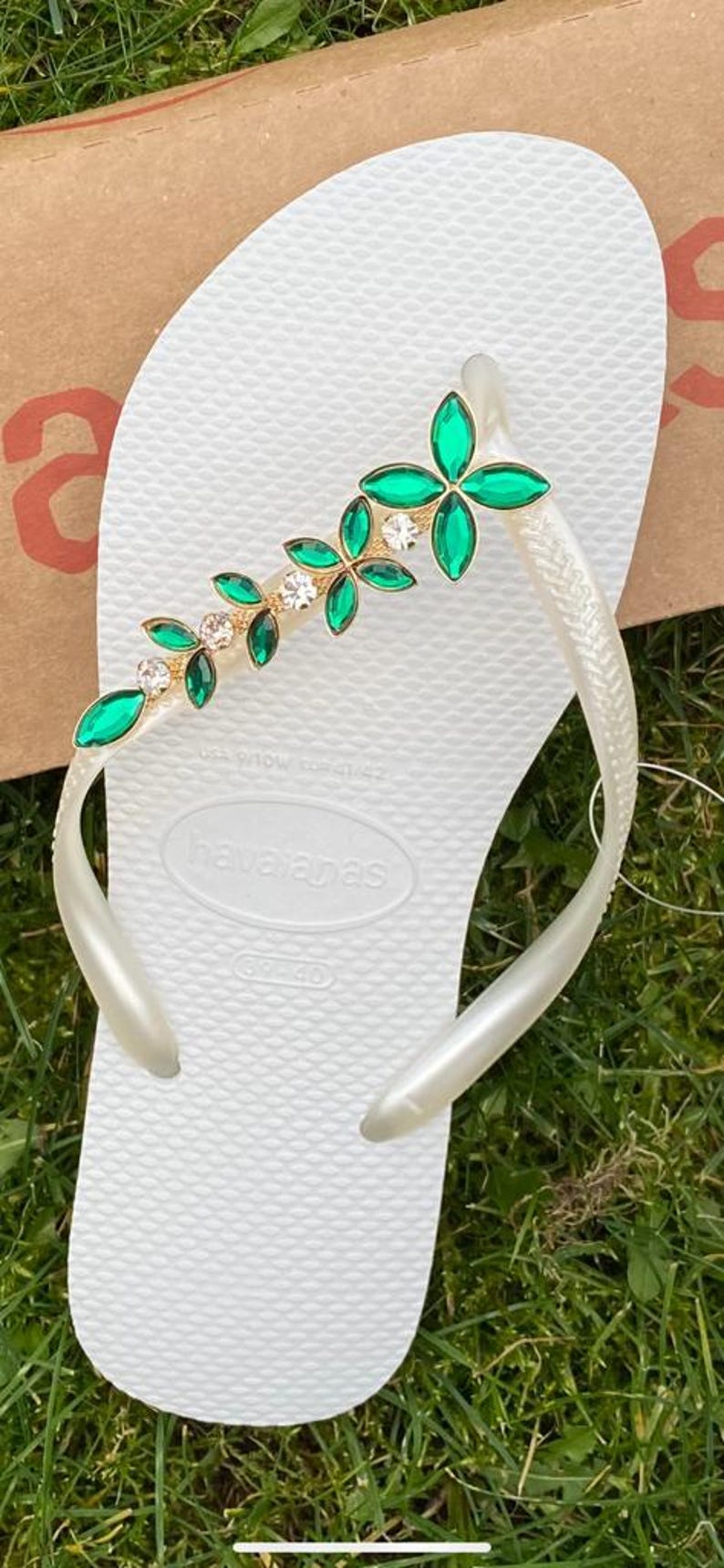 May include: White Havaianas flip-flop with a decorative strap featuring green and clear gemstones in a floral design. The flip-flop is on a grassy surface.