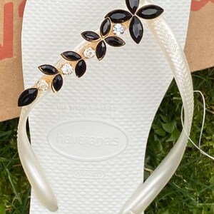 May include: White Havaianas flip-flop with a decorative strap featuring black and clear gemstones. The strap has a gold-coloured base and a floral design. The flip-flop is on a green grassy surface.