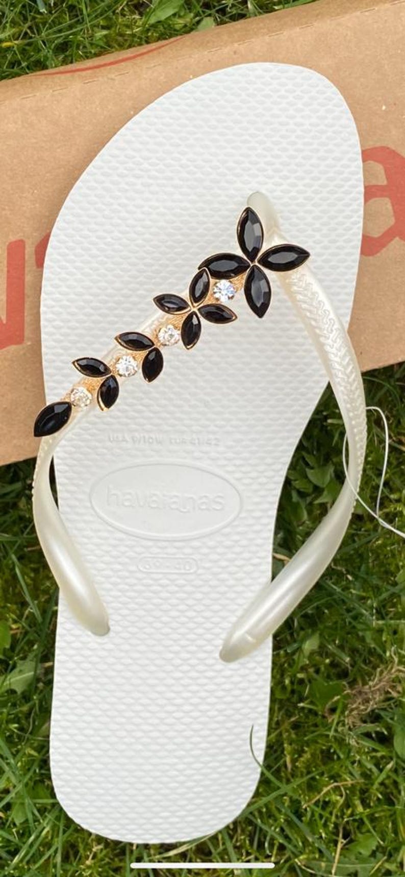 May include: White Havaianas flip-flop with a decorative strap featuring black and clear gemstones. The strap has a gold-coloured base and a floral design. The flip-flop is on a green grassy surface.